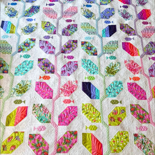 A wide detail shot of a finished Marine Meadows quilt made with the Floral Reef collection, showing elongated hexagon leaf blocks in a vivid rainbow of prints connected by narrow striped branch-like sashing, with small fish shapes scattered throughout the white background.