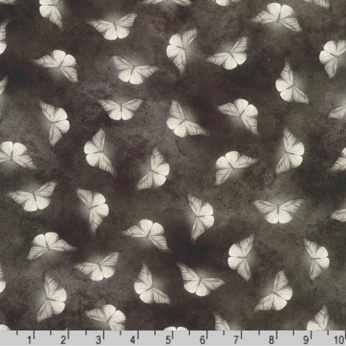 Patterned fabric of softly glowing white moth silhouettes scattered across a dark, mottled charcoal grey background with a ruler at the bottom for scale.