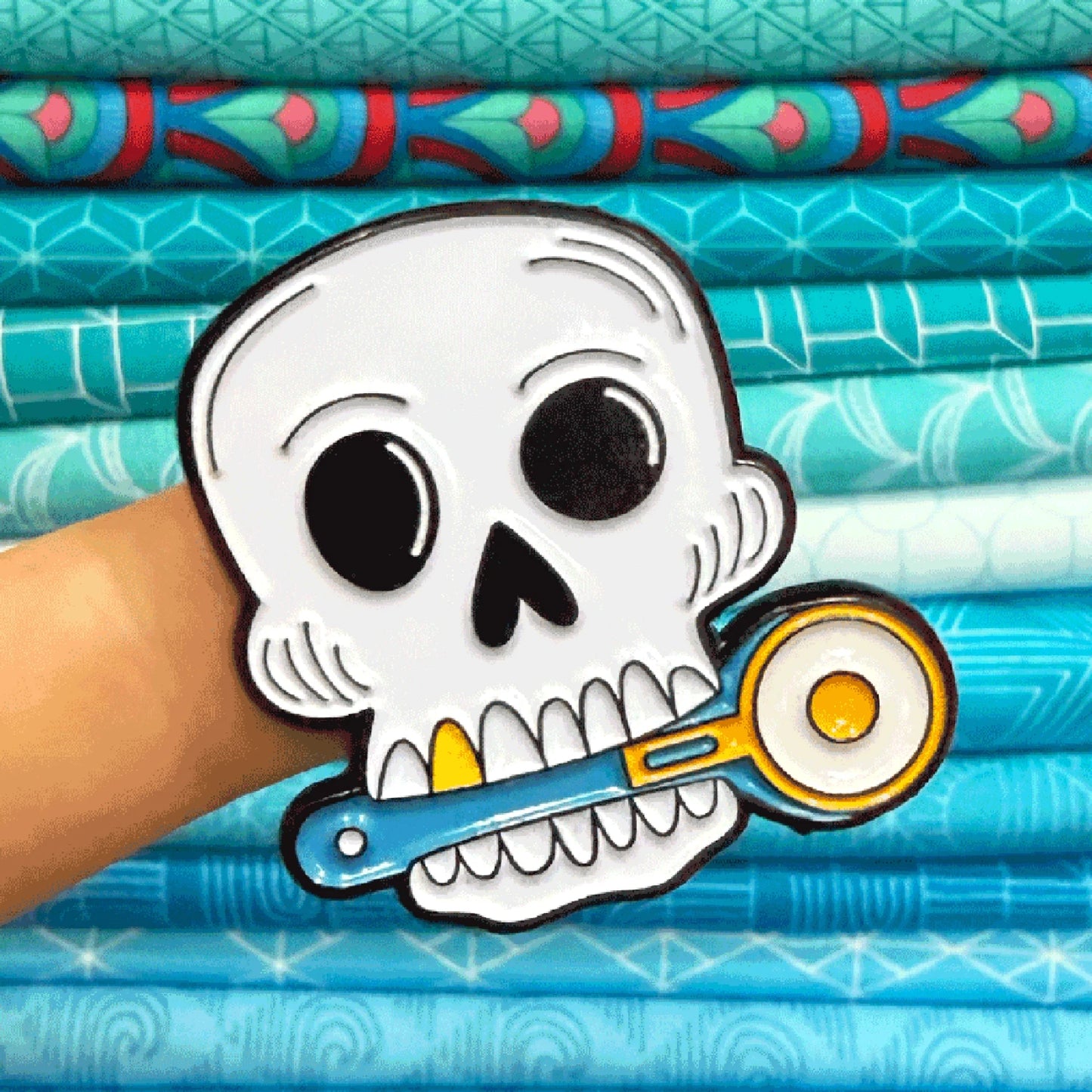 A white skull enamel pin with black outlines holding a blue rotary cutter with yellow accents, held by a hand in front of a stack of blue patterned fabrics.