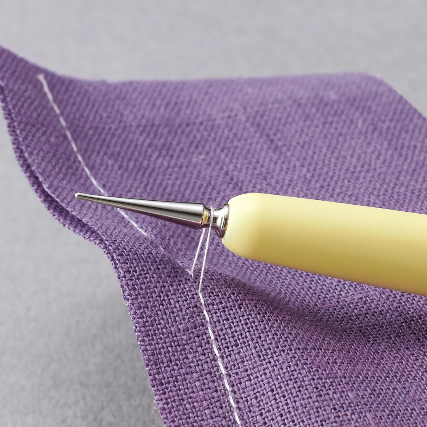 Yellow Hera tool lifting threads from purple fabric with a gray background