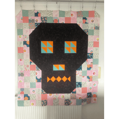 The Bonedigger Quilt features a black and orange pieced skull surrounded by patchwork squares of various pink, green, blue, and grey fabrics. 