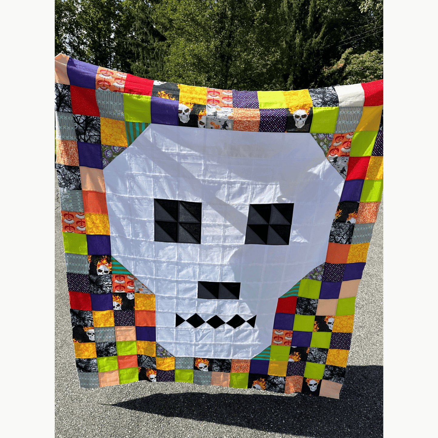 The Bonedigger Quilt features a white pieced skull surrounded by patchwork squares of bright green, purple, orange, black, and yellow scrappy fabrics. 