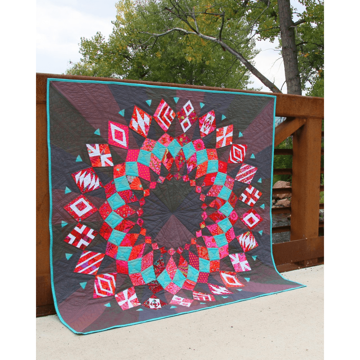 The Cadence Court Quilt made with pink, red, and aqua blue wedges against a dark red and black background.