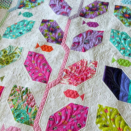 A close-up detail of the Marine Meadows finished quilt showing hexagonal leaf blocks in bold pink, lavender, teal, and chartreuse prints with small fish shapes scattered throughout all set into a cream white background with intricate swirling free-motion quilting.
