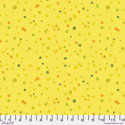 Fabric design of paint droplets in olive, orange, and chartreuse tones scattered across a warm, sunny yellow background. Ruler at the bottom for scale.
