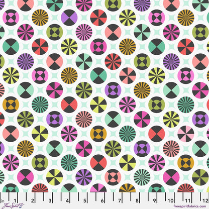 A repeating fabric pattern featuring colorful geometric medallions and pale mint-green star shapes on a white background, with a ruler along the bottom edge showing scale. 