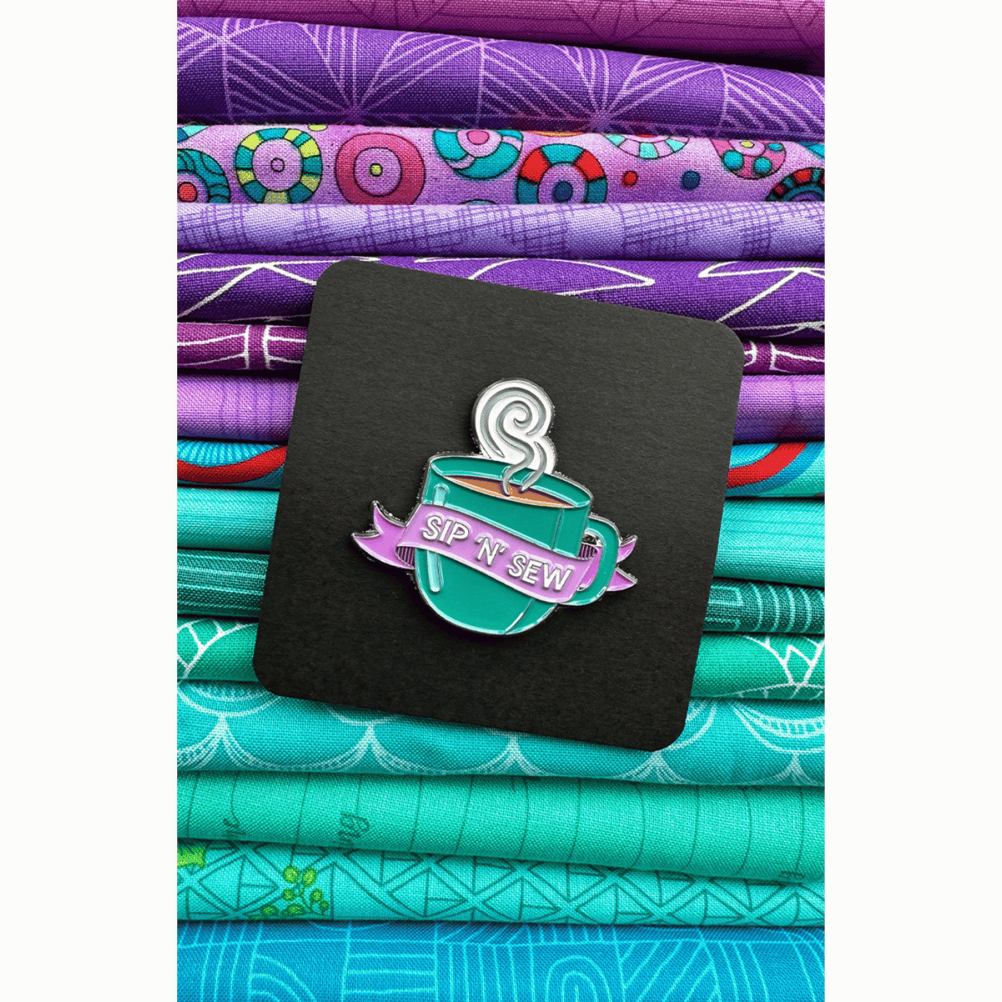 The Sip 'N' Sew Enamel Pin features a green mug full of steaming hot coffee or tea with a purple banner featuring the phrase "Sip 'N' Sew!"