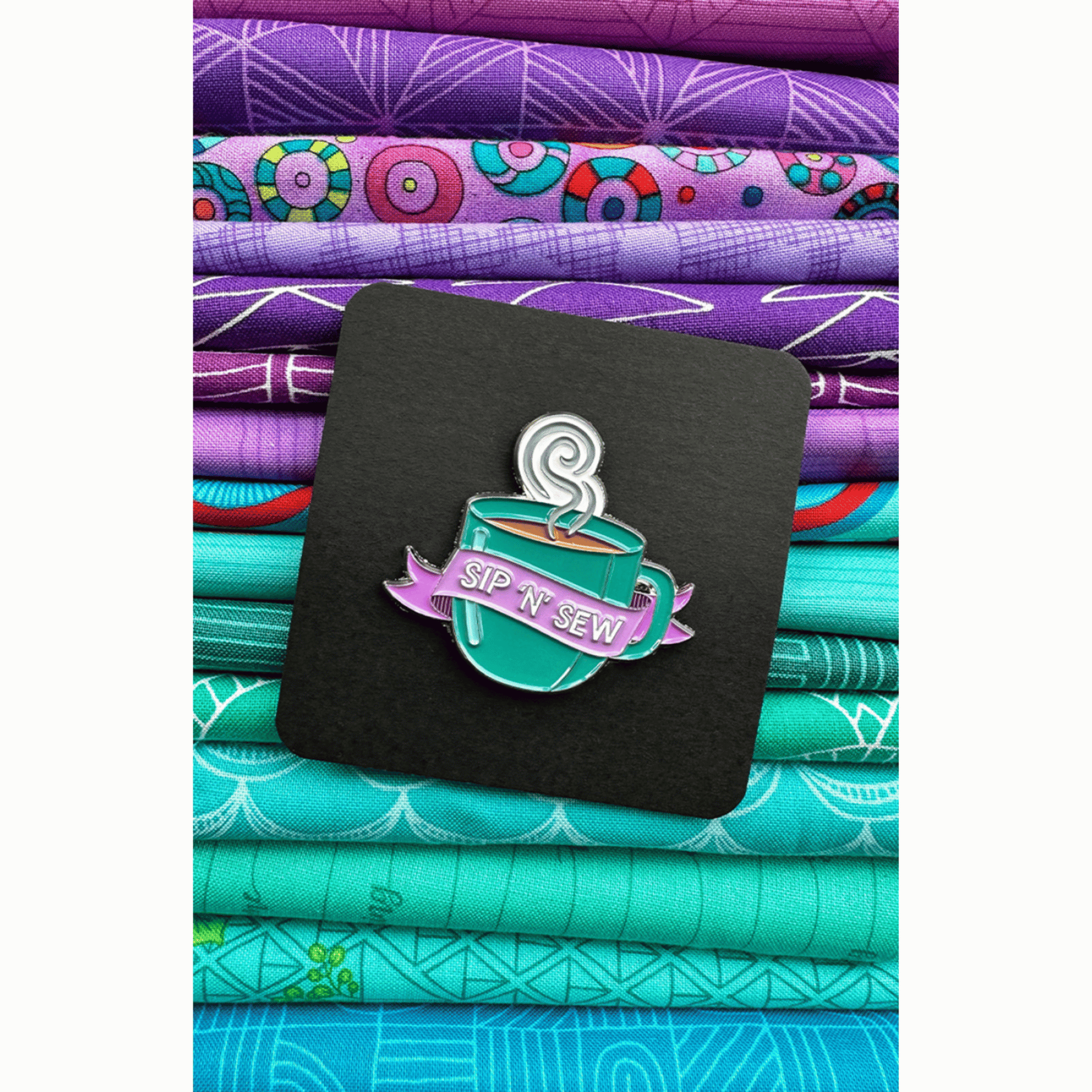 The Sip 'N' Sew Enamel Pin features a green mug full of steaming hot coffee or tea with a purple banner featuring the phrase "Sip 'N' Sew!"