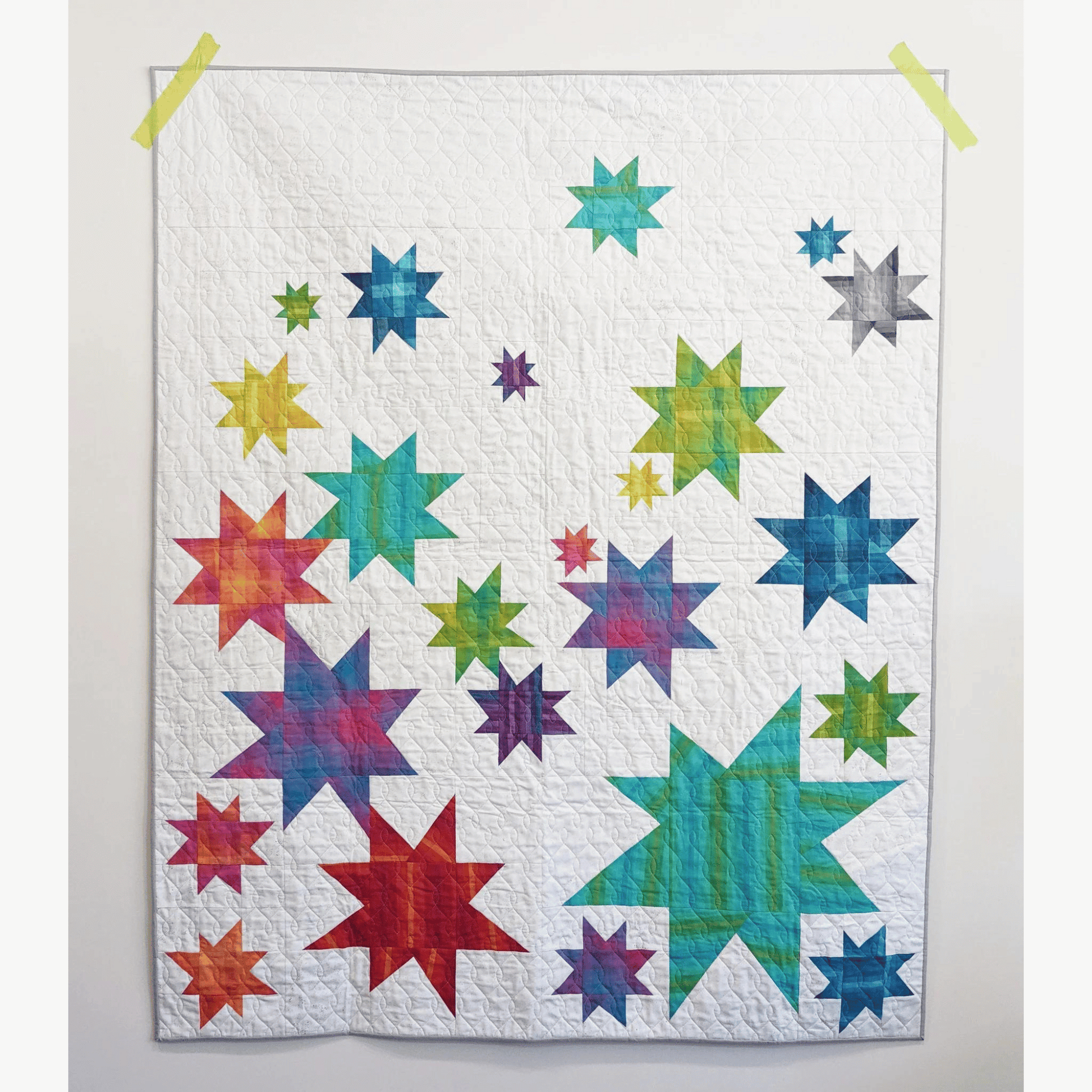 The Vela Quilt is a fun take on wonky stars. This modern quilt features various sized star blocks in blue, green, red, and purple pieced with a white background fabric.